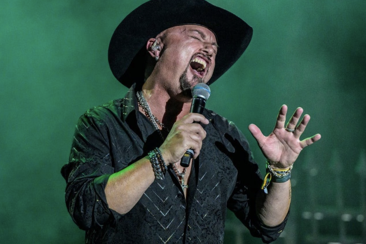 Live performance portrait of former Queensrÿche singer Geoff Tate wearing a black cowboy hat and passionately singing into a microphone on stage.